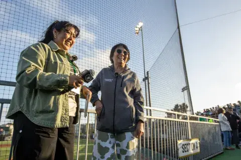 President Ching-Hua Wang of Samuel Merritt University and another woman smile and talk while holding microphones near the baseball field, standing beside the railing in Section A.